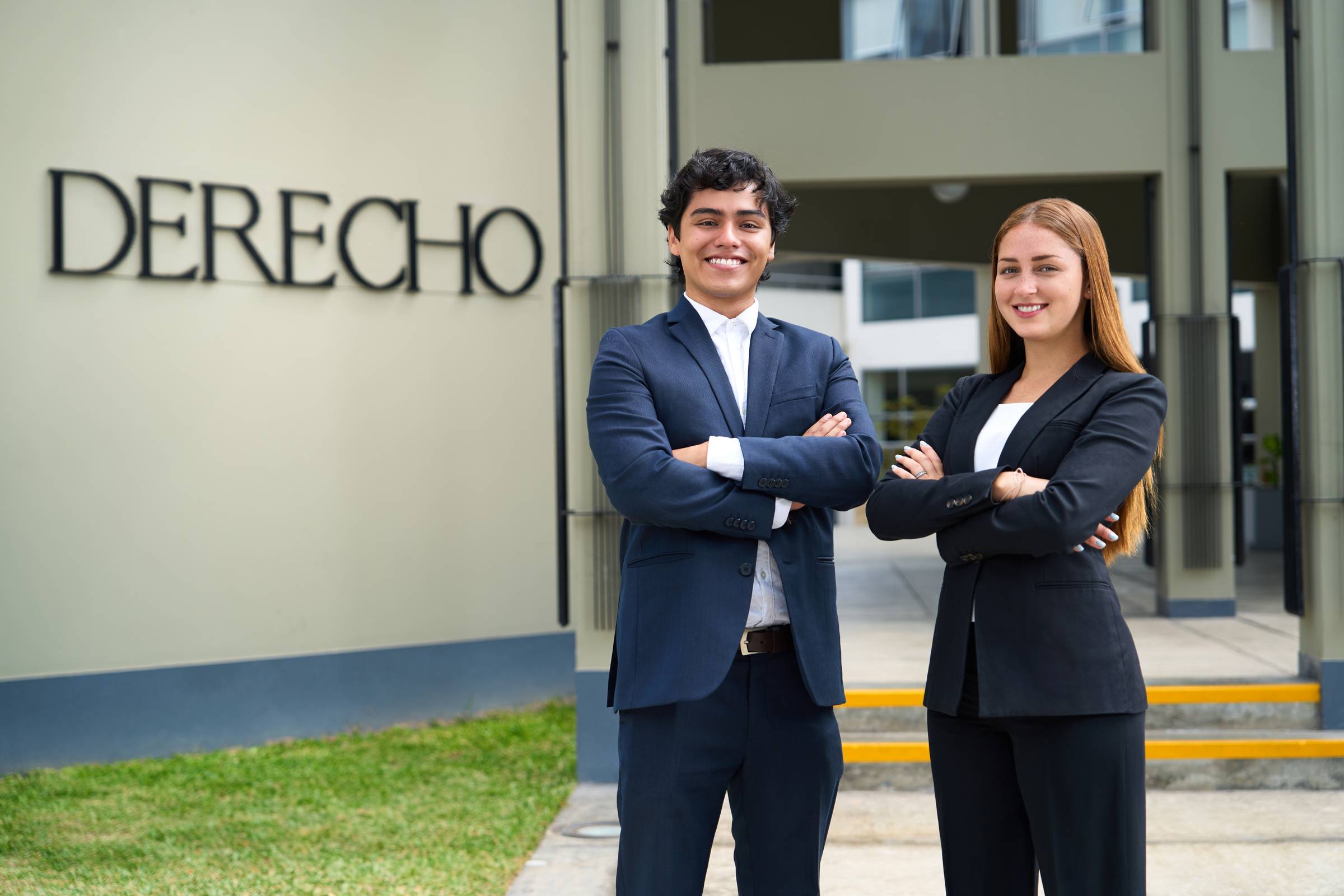 Dos estudiantes de Derecho con traje formal posan con brazos cruzados frente al edificio con el letrero 'DERECHO'.